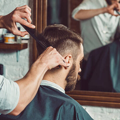 The hands of young barber making haircut of attractive bearded man in barbershop